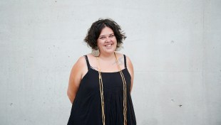 A first nations woman wearing a black dress stands in front of a white background.