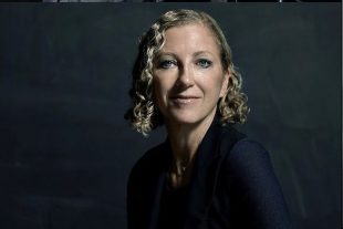 A dark background and a head shot of a Caucasian women with blond shoulder length wavy hair smiling at the camera. She is wearing a dark shirt and has her body turned three-quarters on