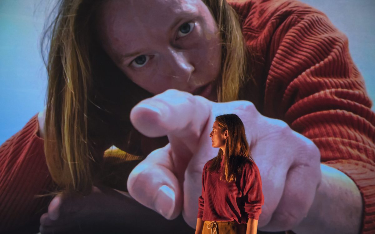 A young woman onstage in front of a large projection of herself.