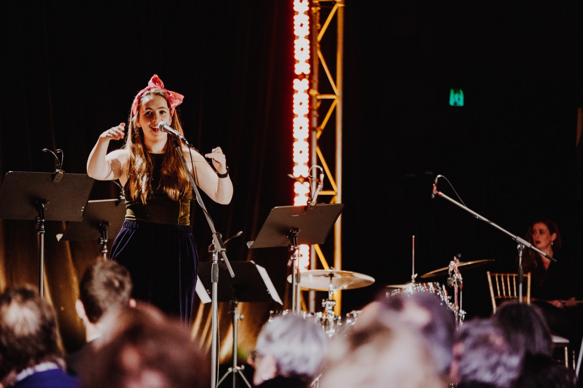 A woman in a reb bow sings into a microphone in front of a crowd.