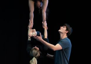 Three circus artists practice a balancing routine in a dark studio.