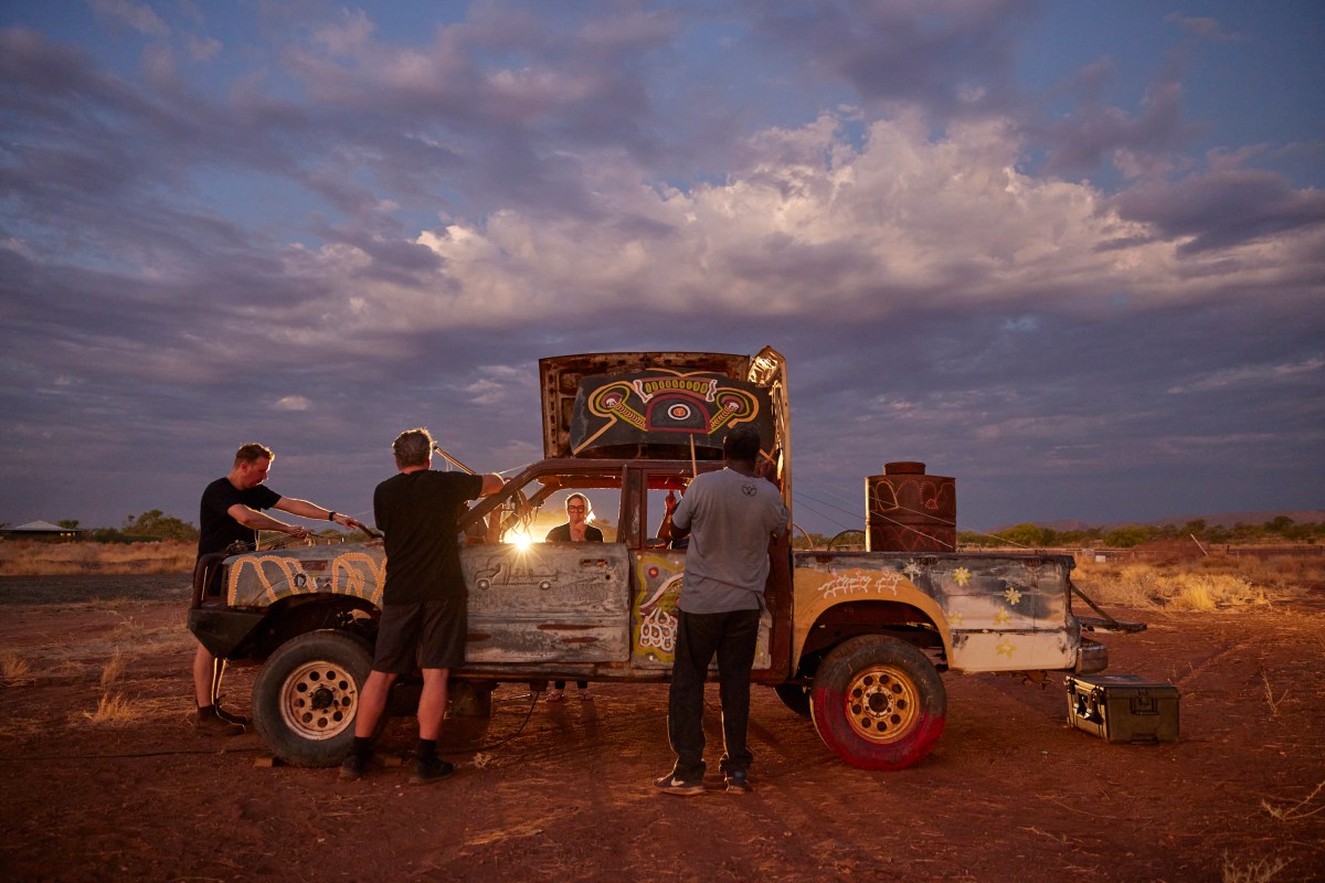 an old car decorated with paint and strings, surrounded by a few people and parked in the desert and sunrise.