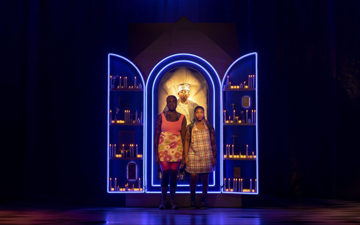 Two young Black women stand on stage in front of an open panel with illuminated blue and gold light. In the centre of the panel is a male figure with gold light illumination around him.