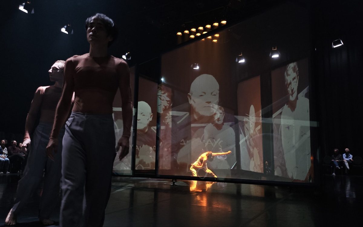 Infinitely Closer. Image is three dancers in tight flesh coloured t-shirtsa and grey pants, two close to the camera, one at the back of the stage, spotlit and bent over, with large screens covered in projections of other dancers. Audience members can be seen seated to the far right and far left.