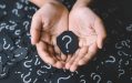 inquiry. Image shows two hands holding a piece of dark card with a chalk question mark on it in front of a black backdrop of chalked question marks.