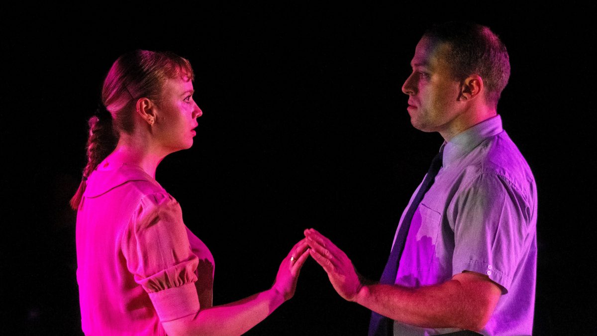 Dead Man's Cell Phone. Image is two actors, one male, one female in a pink light against a black background. They are facing one another and touching their fingertips together.