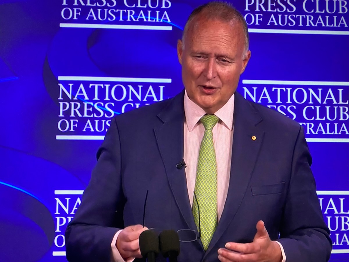 Rupert Myer. Man in suit stands in front of blue wall giving an address.