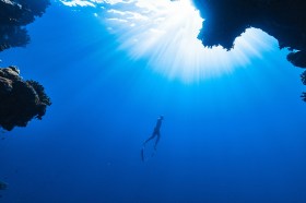 Image is an underwater photo depicting a scuba-diver rising through a deep blue ocean towards the sunlit waters above.