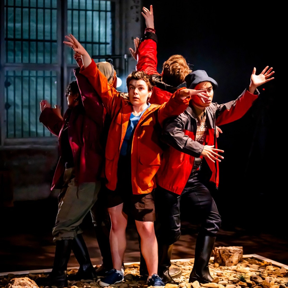 Orlando. Image is of four actors in red jackets back to back with each other and with arms raised in front of them.