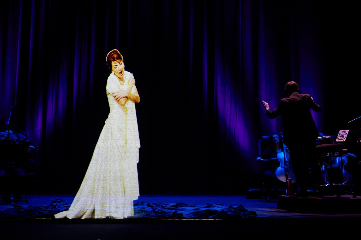 Maria Callas. The image is a holograph of a woman in a floating white dress with dark piled up hair, hugging herself and singing.