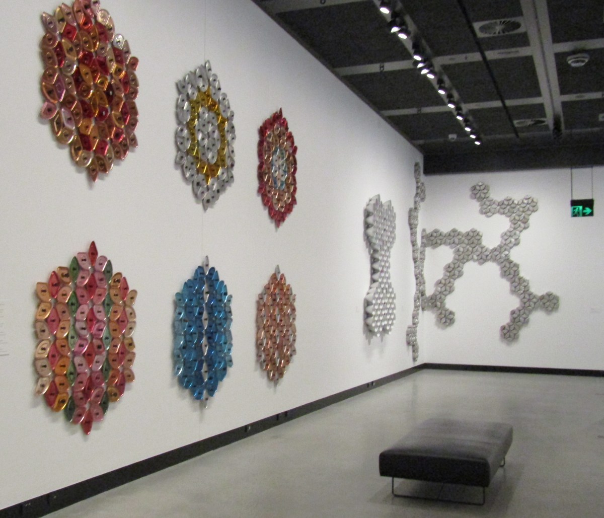 Donna Marcus mesmerises audiences with the mass and variety of vintage cookware she collected, sorted and assembled. 'Radiate' installation view at HOTA. Photo: ArtsHub. Red, blue and silver wall sculptures made of aluminium parts and arranged in different patterns arranged on a white wall.