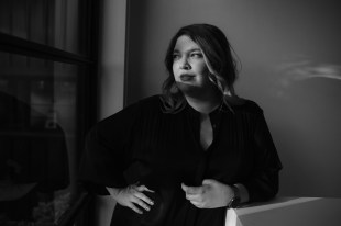 Curator. Image is a black and white shot of a woman with shoulder length hair, and a hand on her hip, wearing a black dress, and looking off to the site. her face is half in shadow.