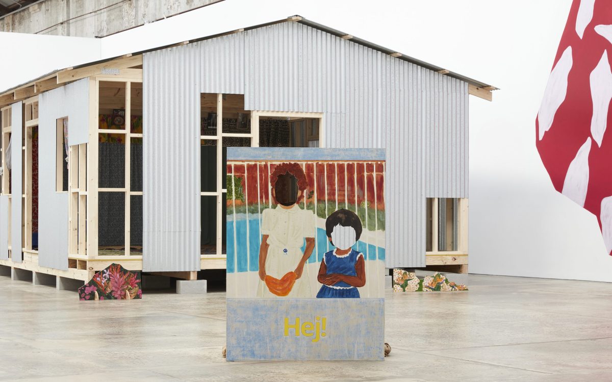 Salote Tawale, ‘I remember you’, installation view at Carriageworks, 2023. Photo: Zan Wimberley. Installation of a Fijian home in a white-cube gallery space. At the front is a free-standing painting that shows two young children with brown skin and their faces hollowed out.