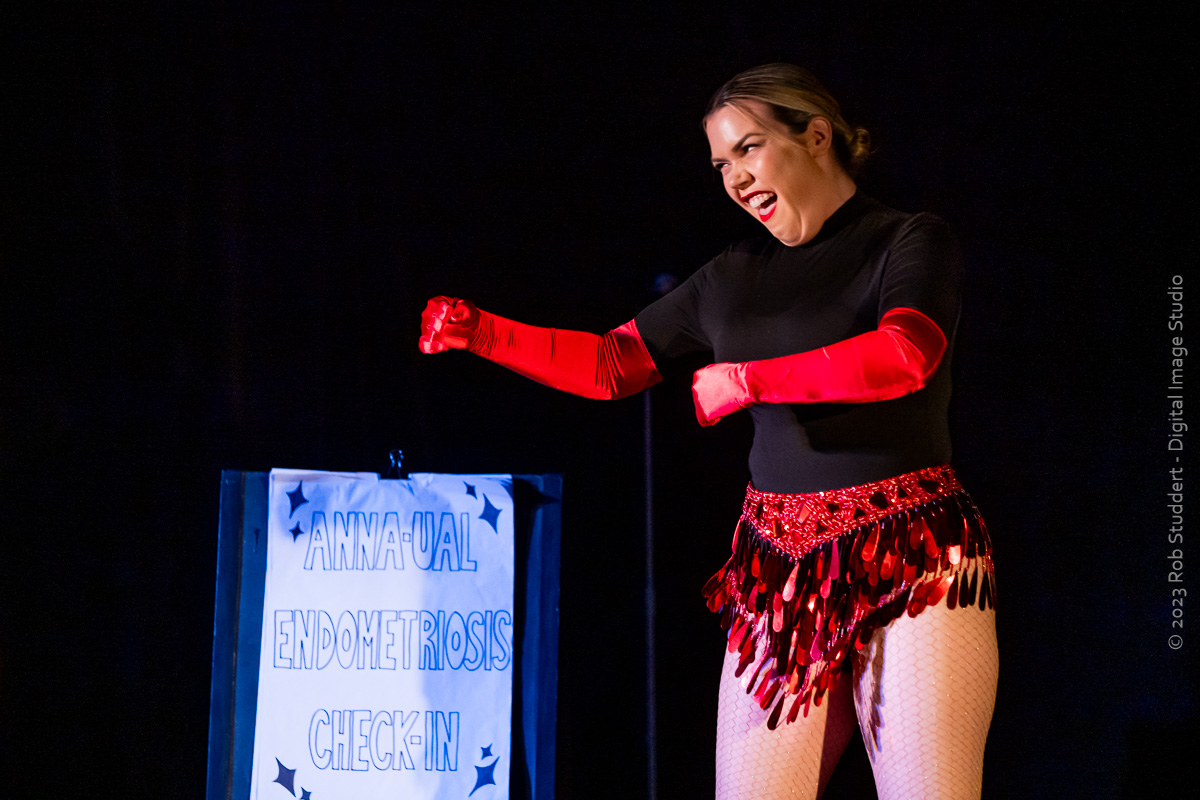 A woman stands on stage against a black backdrop wearing a short black and red tasselled costumer with long red gloves. She is laughing and grimacing and standing next to a sandwich board.