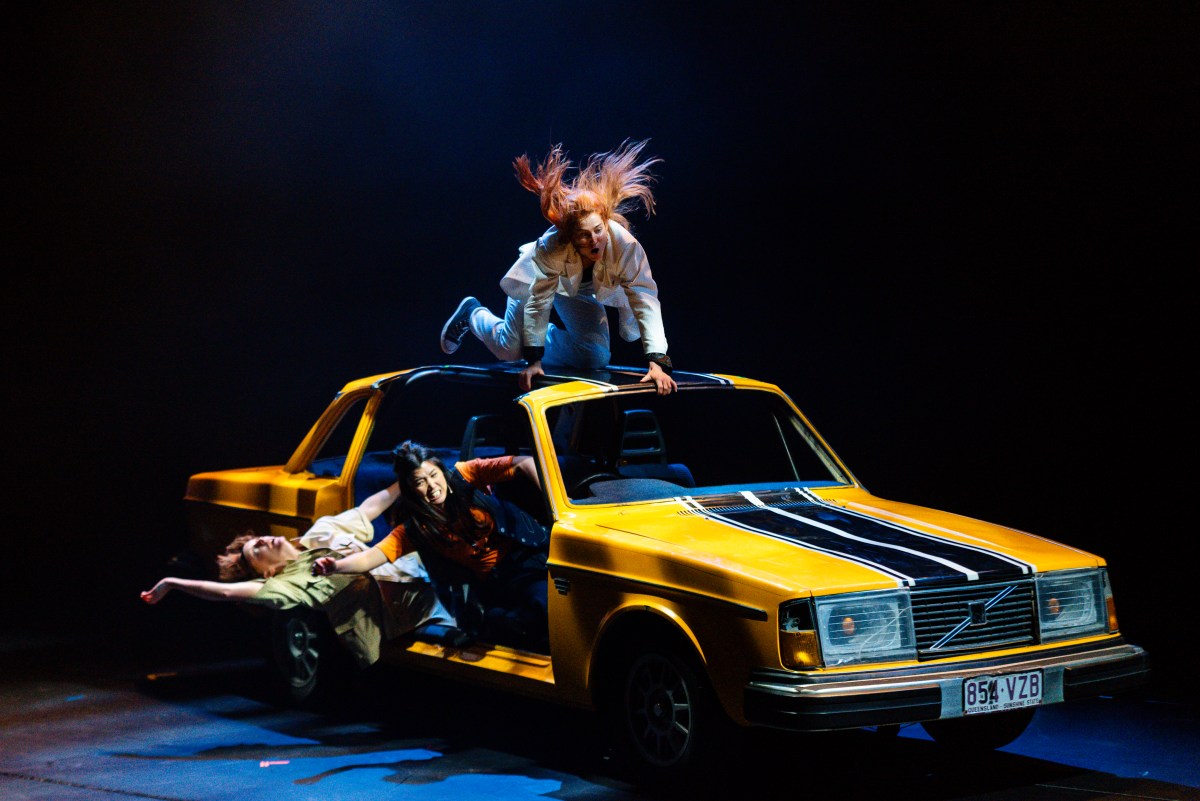 Three people are hanging out of a yellow car, which has cut-off panels. Stunt Double.