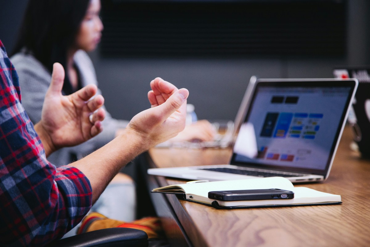 Building creative courses is a great way for many artists to have a side hustle. Image is two people sitting at a desk in front of an open laptop. We can't see much of the first person, other than that they're wearing a red checked shirt and holding their hands out with their palms facing them.