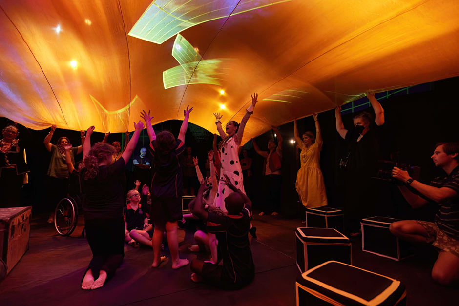 A group of children lead by a facilitator in the middle standing beneath a large orange sheet with green projections. A couple of adults are holding up the sheet, while the kids have their hands raised in the air. 