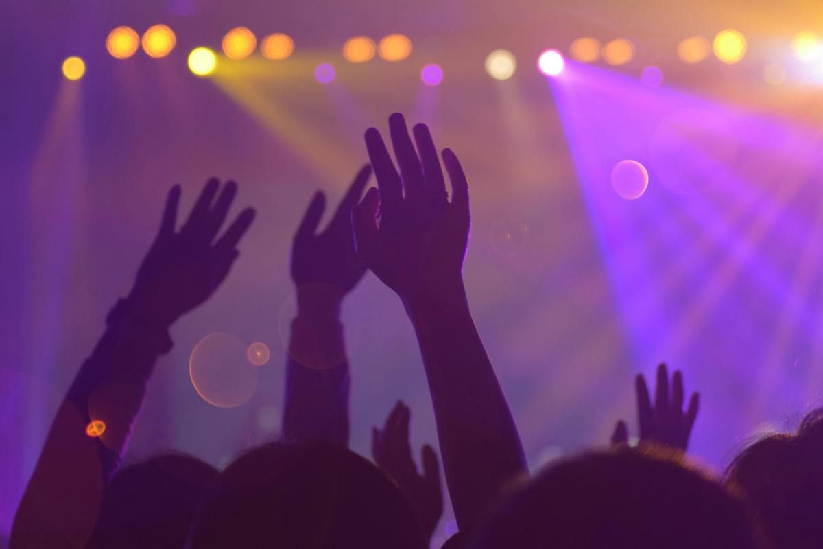 A concert venue with three raised hands reaching upwards fro the crowd, through purple and yellow spotlights. Wellness comedy.