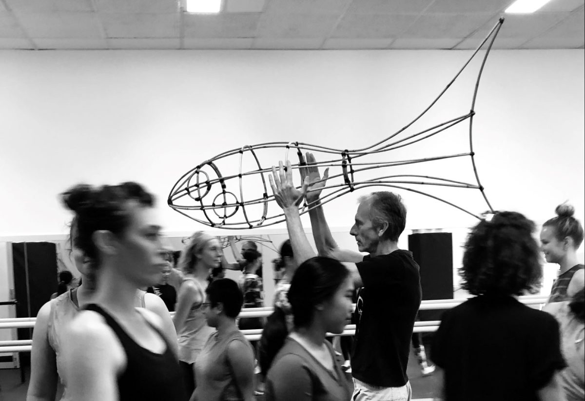 Tim Newth exit interview. A black and white photo of a diverse group of performers in a rehearsal room. Tim Newth, a older man, stands at the rear holding a wire-framed fish prop over his head.