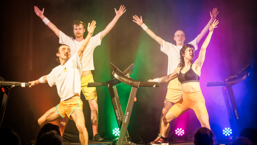 Four performers on treadmills throw their arms up in the air and are spotlit from behind by green, blue and purple lights.