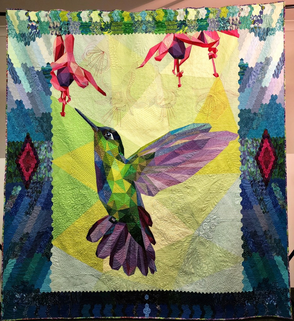 ‘The Honeycombed Hummer’ by Lyn Crump for the Australasian Quilt Convention. Photo: Supplied. A detail quilt with vibrant colour blocs featuring a realistic humming bird at the centre in mid-flight, surrounded by several red and purple blooms. Crump speaks on the cost of her craft. 