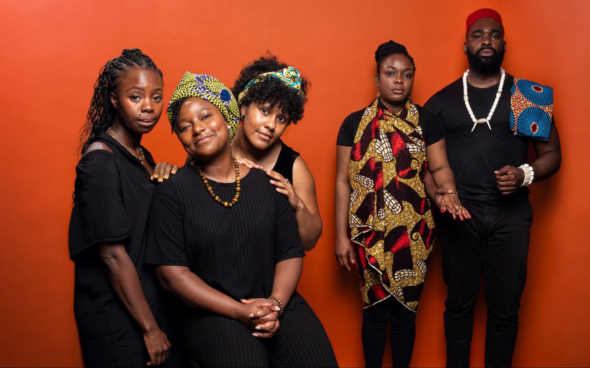 Members of the cast from ‘Akaraka’ by Amarachi and produced by Empty Seat Theatre. Photo: Cameron Grant. Two people standing close to each other with their hands on the person seated in front of them, smiling. Two more people are standing at the back and off to the right hand side, their hands are held together looking slightly serious. All members of the cast have dark skin brown skin and black hair, with a red-orange background.