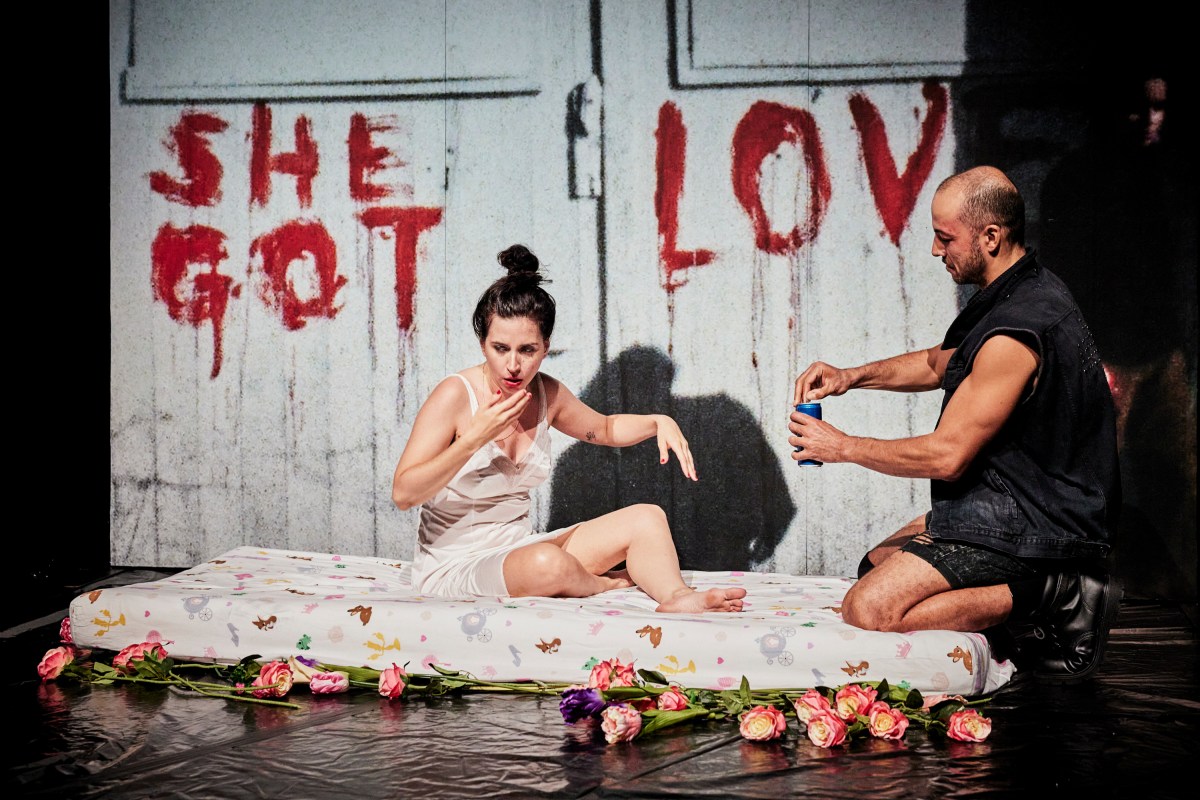 RISING. A woman in a slip sits on a mattress on the floor, while a man at the foot opens a can of drink. Behind the mattress is a panel with the words SHE GOT LOVE daubed on in red paint.