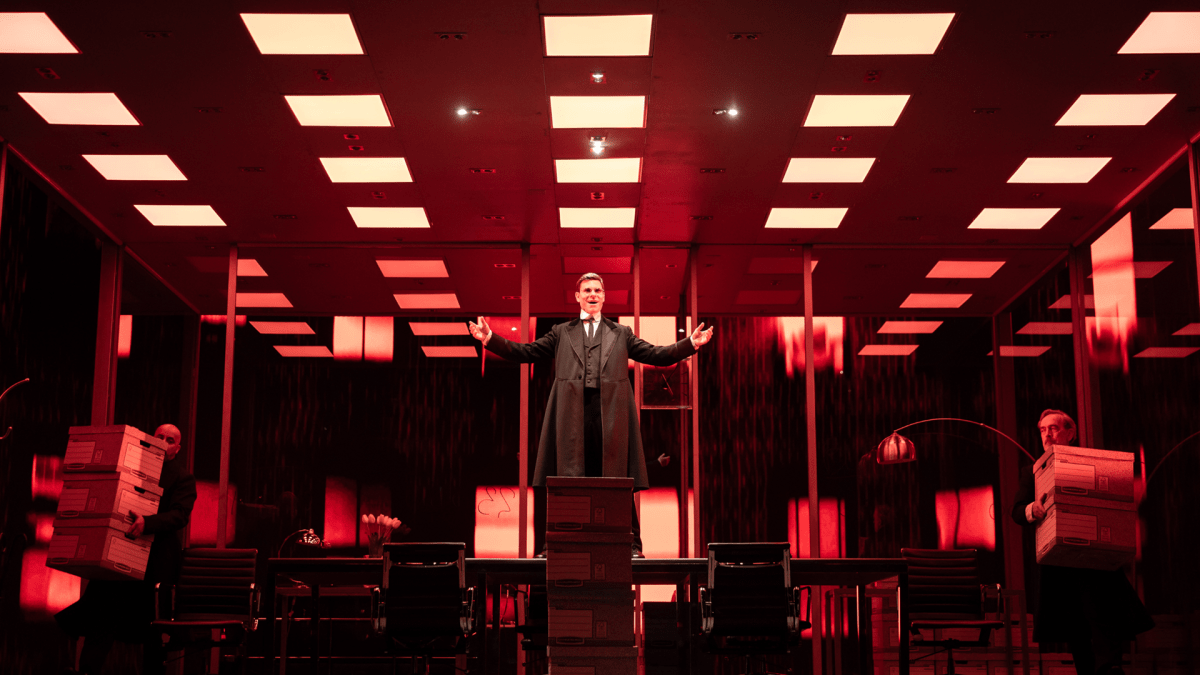 Lehmann Trilogy. Image is a man in a red tinted corporate office, with a grid of square fluorescent lights overhead. He is standing on a boardroom table with his arms outstretched to the side. Around him are file boxes and one is being carried out by an office worker.