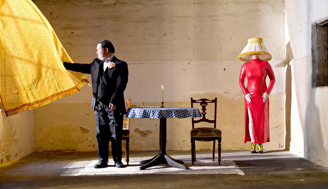 Private View. Image is of a man with Down's syndrome standing in a room looking out of a window and holding a billowing yellow curtain. In the middle of the room is a smalll round table with a lit candle on it and two chairs. In the right hand top corner is a female figure in a slinky figure hugging red dress, with a lampshade obscuring her face and head.