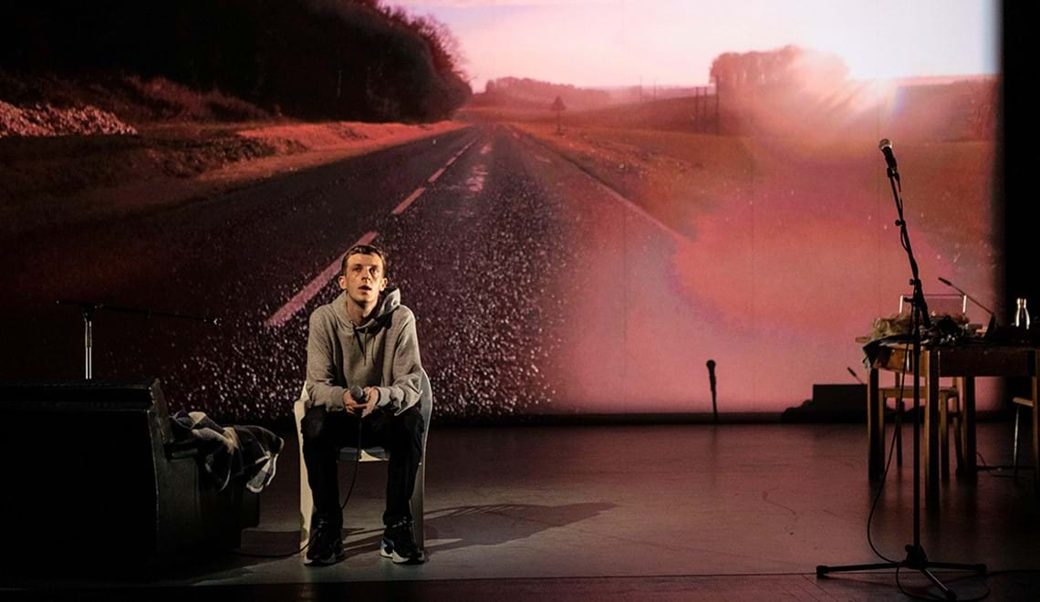 Qui a tué mon père. A man sits on a stage in front of a backdrop projection of an open road.