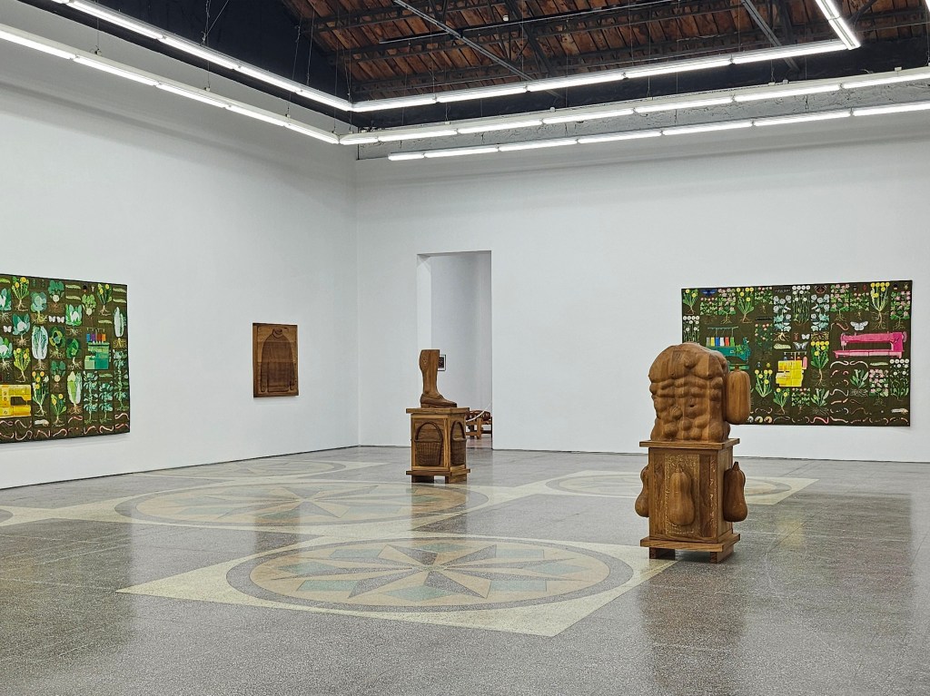 ‘The weaver and the spoke’, installation view at Antenna Space. Photo: ArtsHub. A spacious gallery with white walls and marble patterned floor with two sculptural works on display and a series of textile works. The sculptors are wood carved and depict aspects of the human body combined with agriculture harvest such as pumpkins. The tapestries depict sewing machines among the farm land. 
