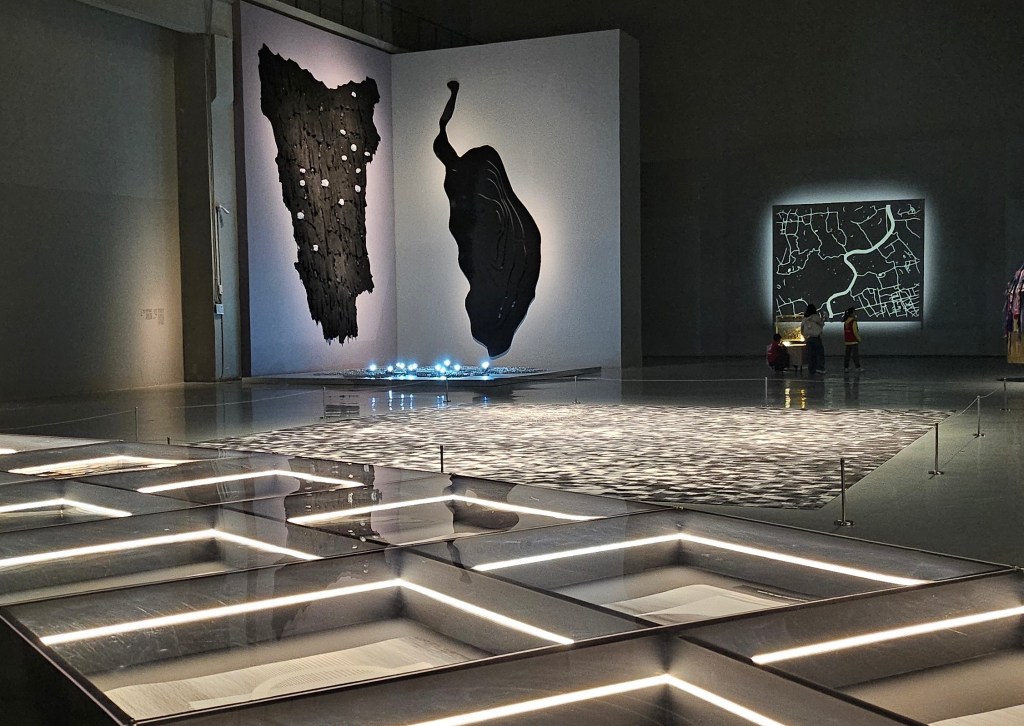 ‘14th Shanghai Biennale: Cosmos Cinema’, installation view at Power Station of Art. Photo: ArtsHub. Installations inside a grey concrete space with LED lights. On the far corner is a black textile work shaped like geographical maps. 
