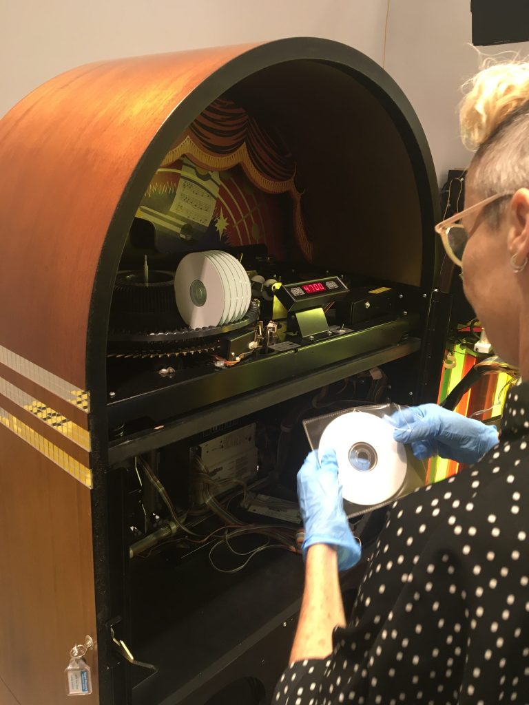 Lisa Mansfield working on the jukebox by artist Susan Hiller, ‘Die gedanken sind frei (Thoughts are free)’, 2012 for ‘What Does the Jukebox Dream Of?’ at AGNSW. Photo: Supplied. A person with a blonde pixie cut wearing glasses and a black shirt with white dots removing a small white CD from its plastic package. In front of them is the inner compartment of a jukebox. 