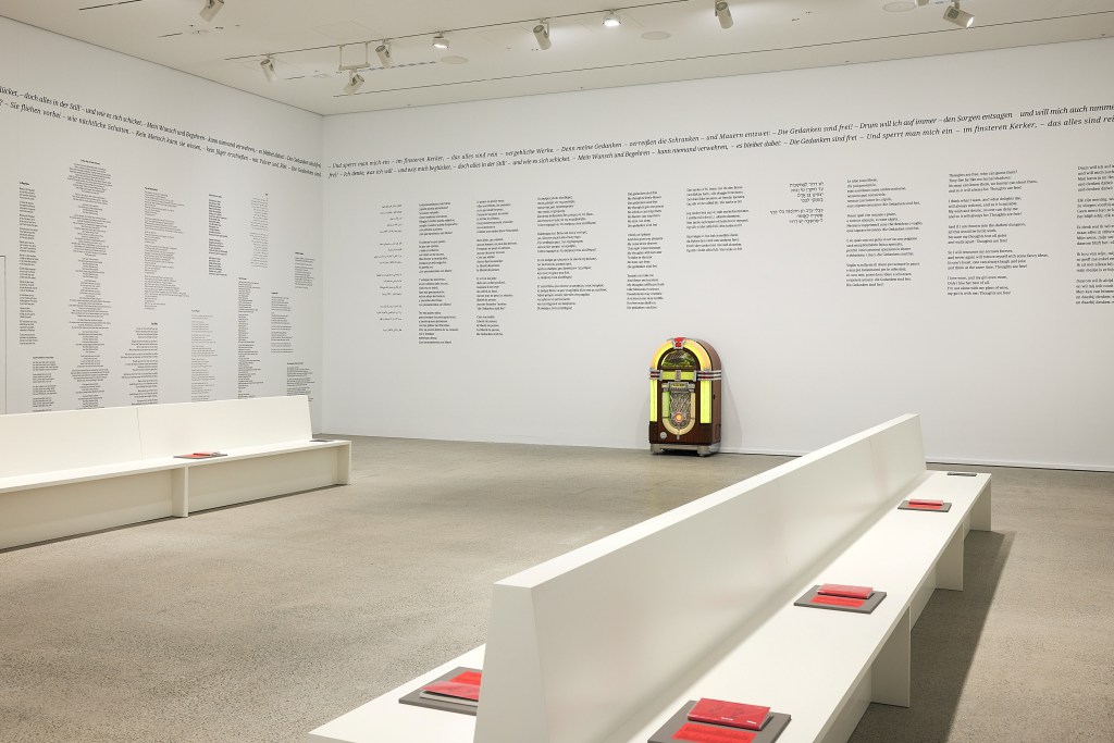 ‘What Does a Jukebox Dream Of?’, installation view at AGNSW. Photo: Supplied. A single jukebox with yellow neon lights stand inside a white-walled gallery space with concrete floor and two rows of white benches. On the walls are rows of (illegible) text, likely to do with the songs in the jukebox. On each bench are several booklets with a red cover. 