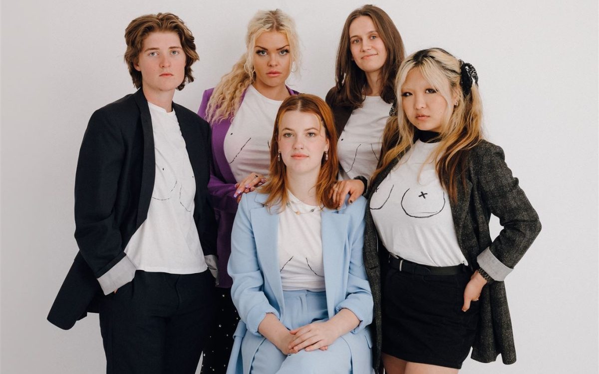 Four female and non-binary people stand in a group facing the camera, one is sitting at the front wearing light blue, the others are standing behind her. All five wear a T shirt with breasts and crosses for nipples drawn on in texta.