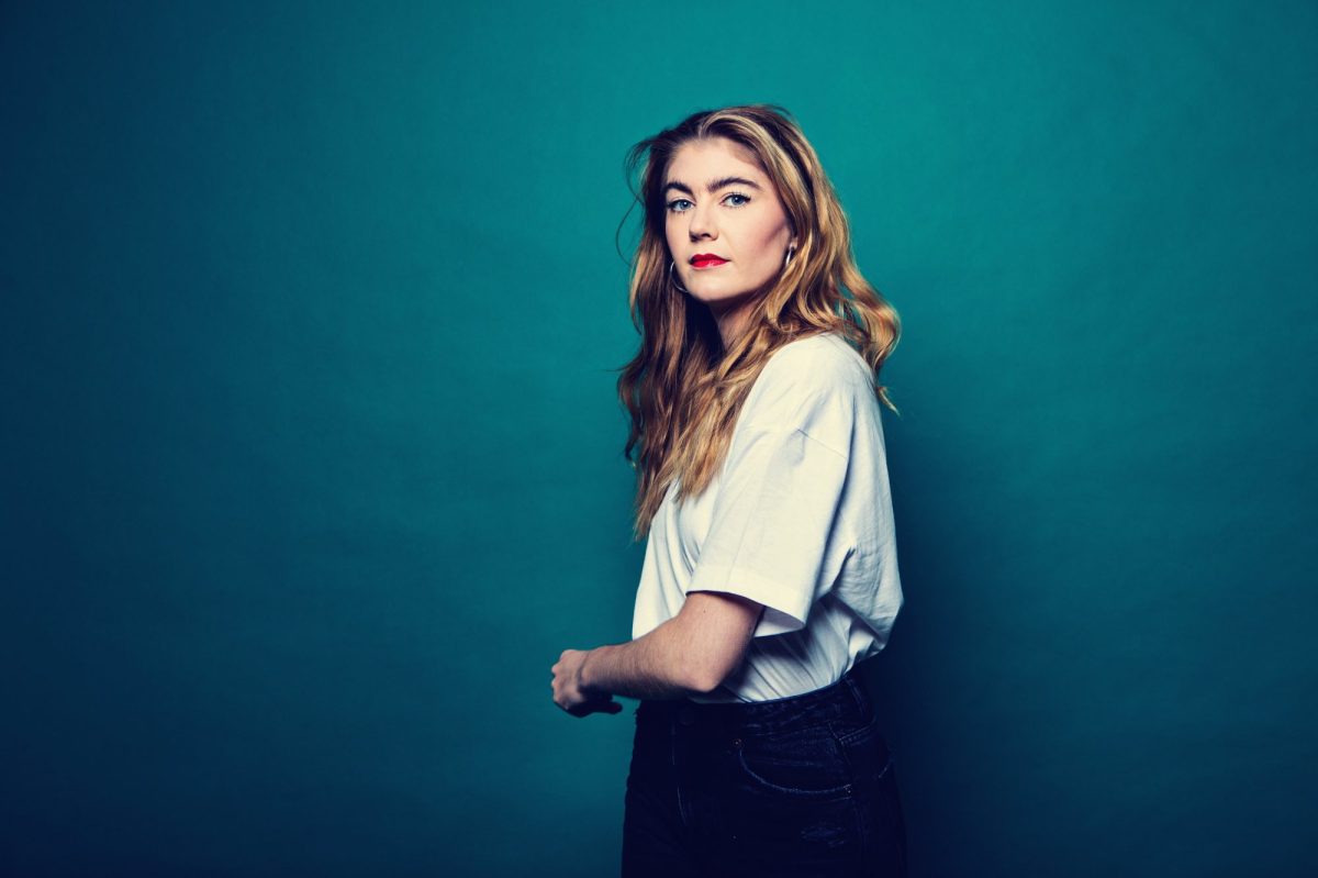 A young white woman stands side on against a greeny/blue backdrop. She has long wavy light brown hair, bright red lipstick and is wearing a white short sleeved shirt and black pants. Brodi Snook.