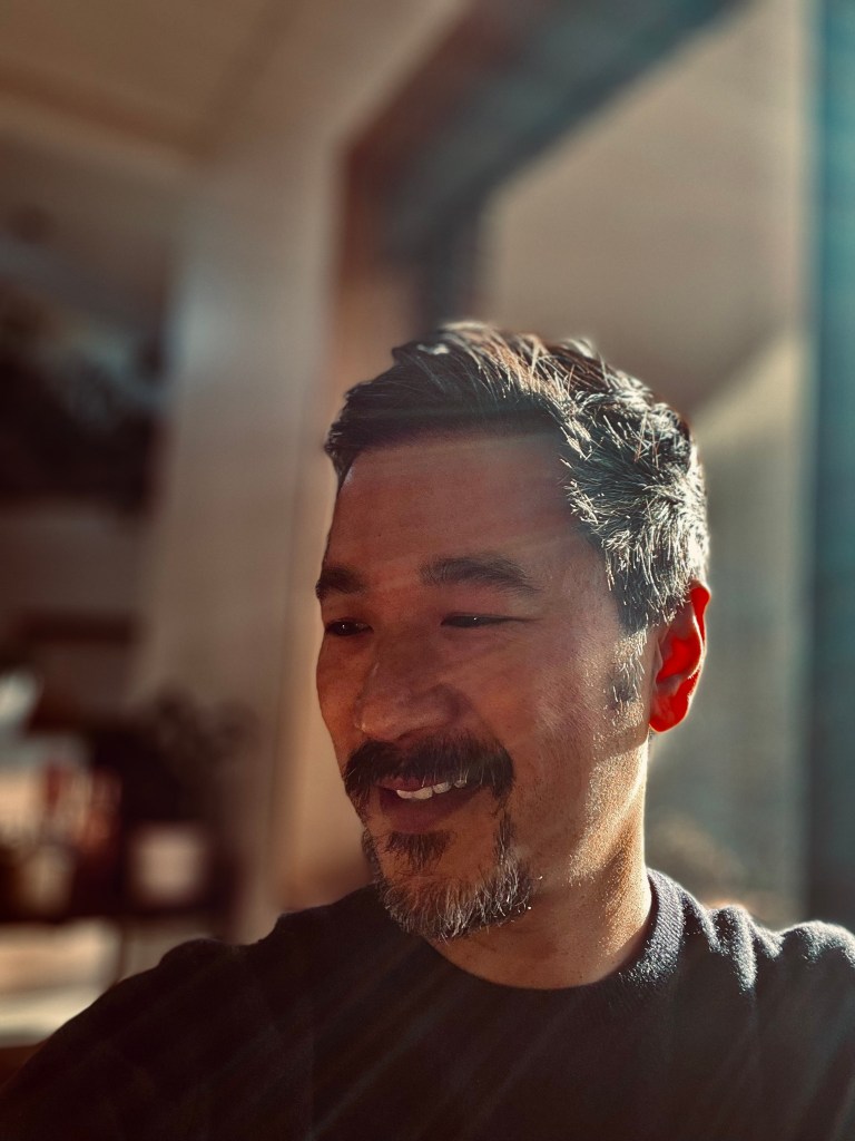 Poetry. Image is a man of Vietnamese appearance with a moustache and goatee beard, standing in front of a window so the light streams across the side of his face. He is looking off to the left and smiling.