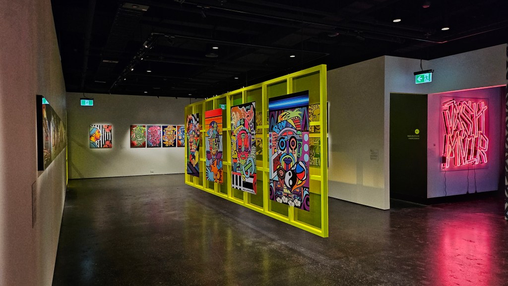 'JXSH MVIR: Forever I Live', installation view at Koorie Heritage Trust. Photo: ArtsHub. Colourful works in a gallery space, featuring a neon yellow panel with digital illustrations in the middle, and a neon light spelling 'JXSH MVIR' on the right hand side.