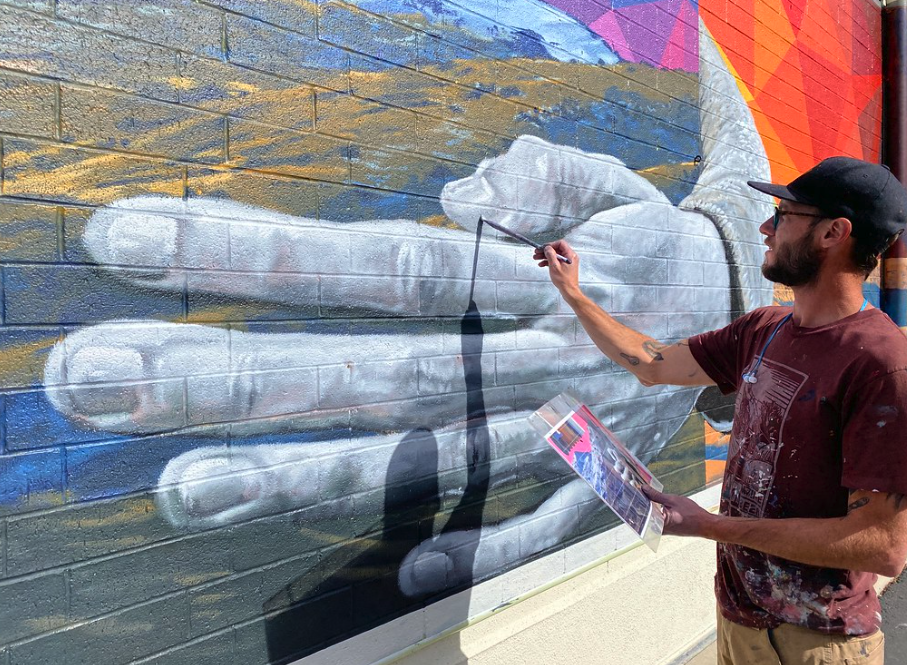 Nevada murals. A close-up of a giant hand being painted in a mural by a Caucasian man in a baseball cap.