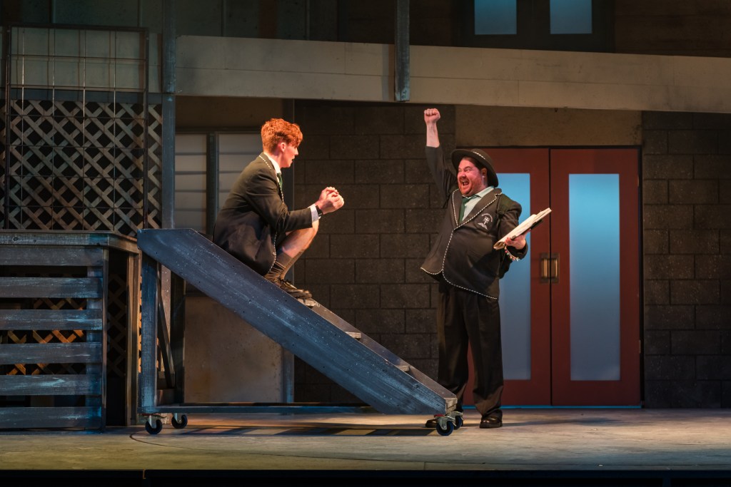 Fourteen. A stage set in which a young red haired boy in school uniform sits on some wooden steps facing the right. In front of him stands a grown man also in school uniform raising his right hand triumphantly.