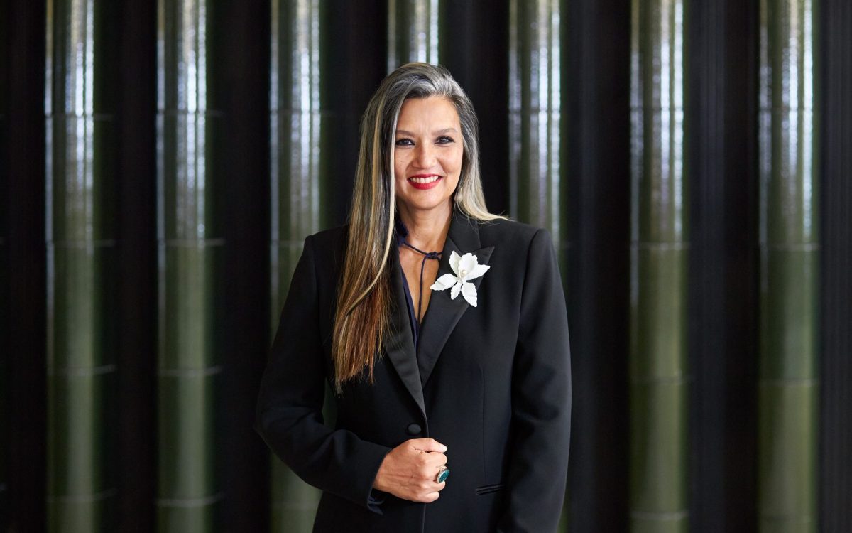 Suhanya Raffel, Museum Director, M+. Photo: Winnie Yeung @ Visual Voices. Image: Courtesy M+, Hong Kong. Raffel against a green tiled architectural background. She is a middle-aged woman with long black and grey hair, wearing red lipstick and smiling at the camera. She is dressed in a black blazer with a white flower brooch.