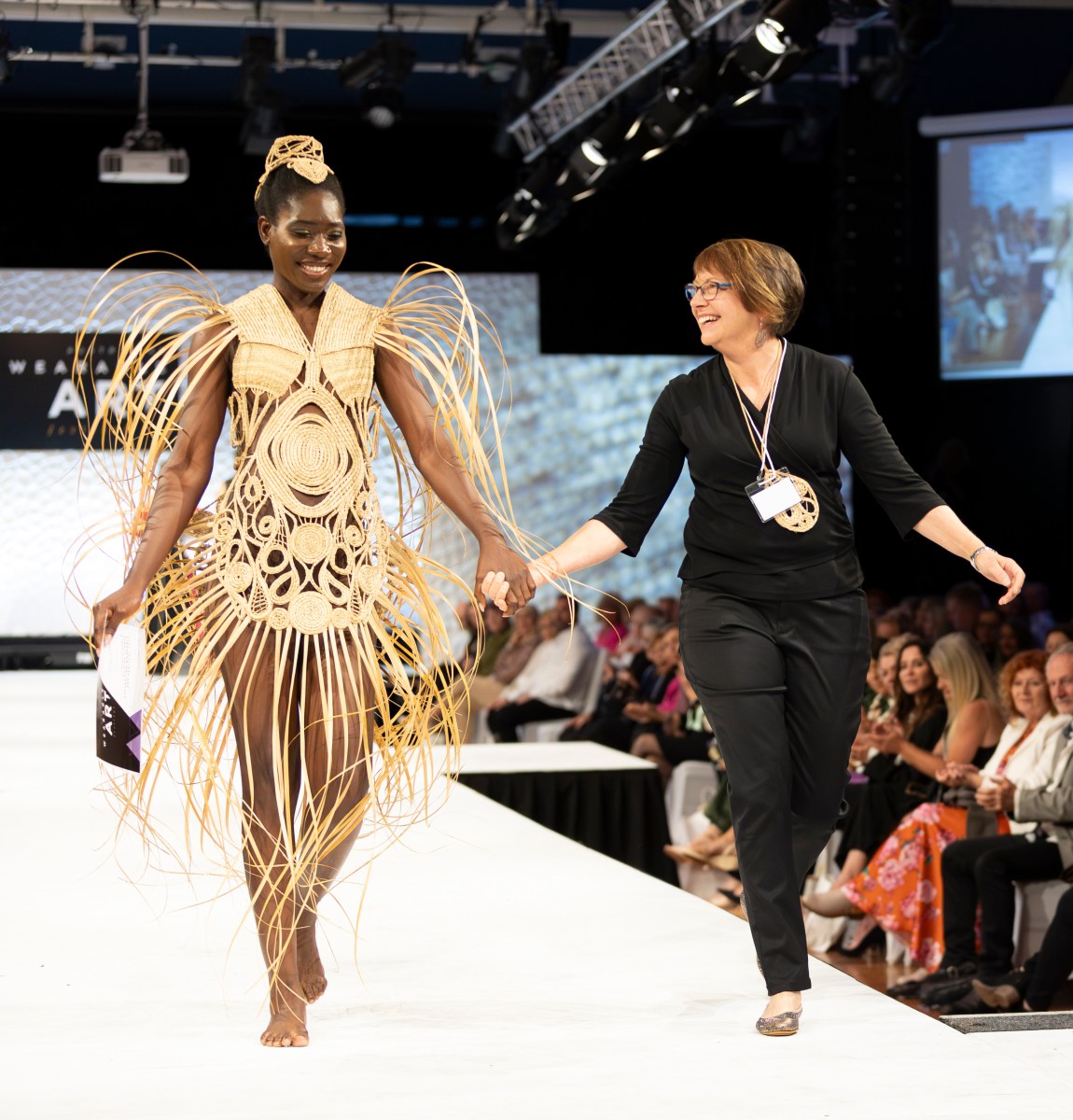 A woman walks down a catwalk in an elaborate costume made out of woven yellow reeds. She is holding hands with a designer who is all in black and smiles at her.