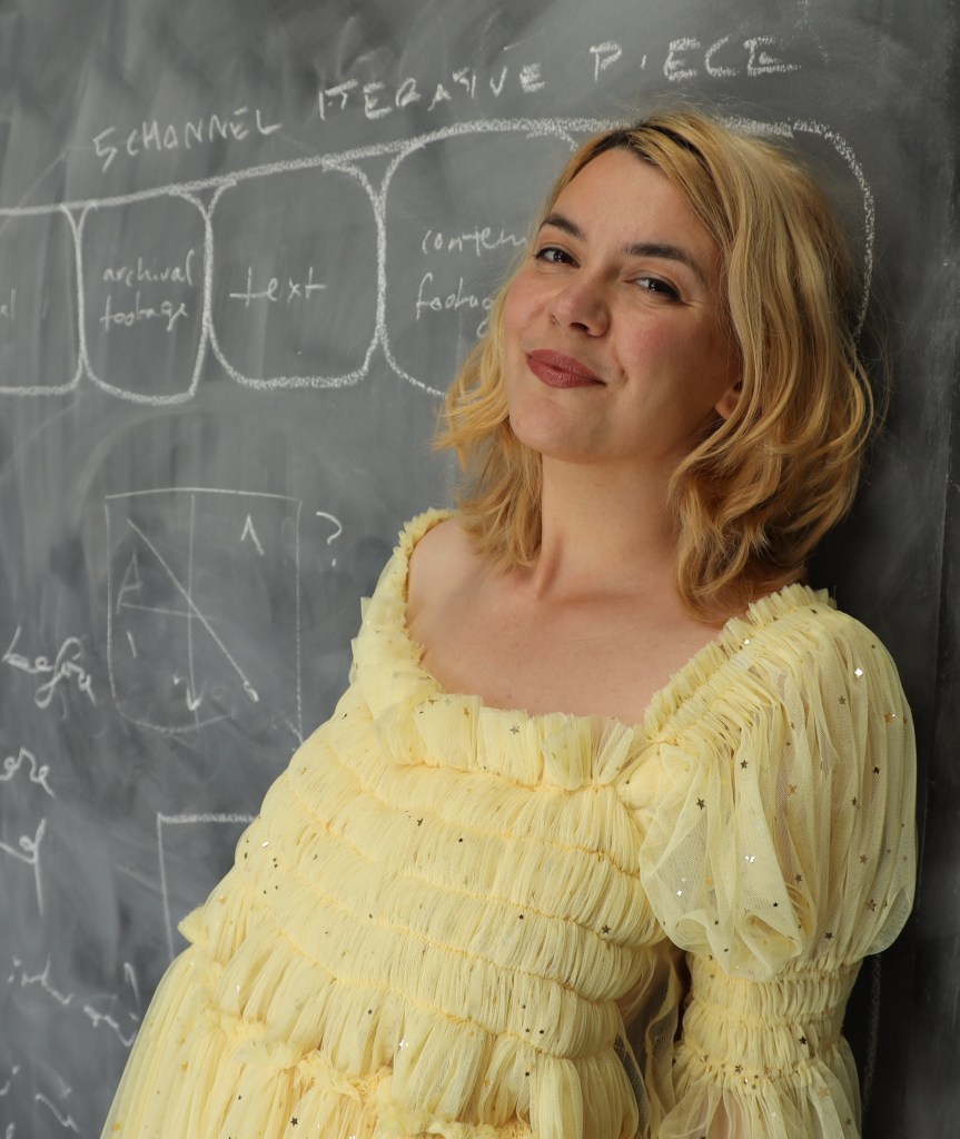 Poetry. A fair skinned woman with blonde hair and a yellow ruched dress leans against a blackboard and smiles at the camera.