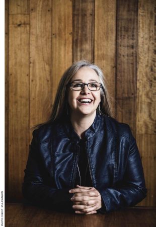 Hidden Thoughts. MSO. A white woman with long straight greying hair and glasses is looking slightly up and to the right with a big smile. She stands in front of a wall covered in wooden boards and wears a leather jacket.