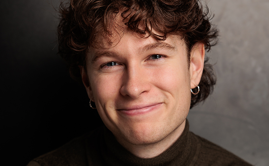 A headshot of a young man with wavy brown hair and a black top. He is grinning