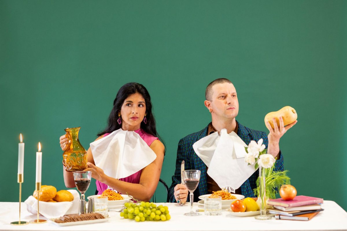 Peacemongers. Against a green backdrop an Asian woman and white man sit at a dinner table laden with food, candles and books. He holds a butternut pumpkin and she holds a wine glass. They are both wearing serviettes tucked into their collars.