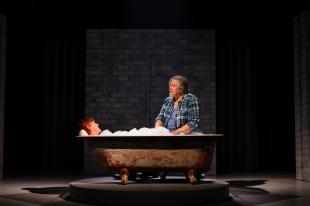 A woman is lying in a bathtub covered in bubbles. A man in a blue checkered shirt and t shirt sits nearby her and looks down at her.