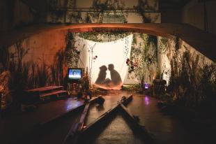 The silhouette of a woman and a man can be seen behind a lit up sheet. Around them there are planks of wood, greenery and a TV set.