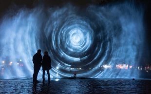 ‘Constellations’ by Joanie Lemercier to feature at Now or Never 2024. Photo: Supplied. A large projected vortex glows above the water at night. Two people are standing close to the artwork.