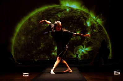 A dancer stands posed, arms outstretched and slightly crouched. She holds an arrow in her teeth. Behind her is a dramatic, green-tinted photograph of the sun depicting several solar flares.